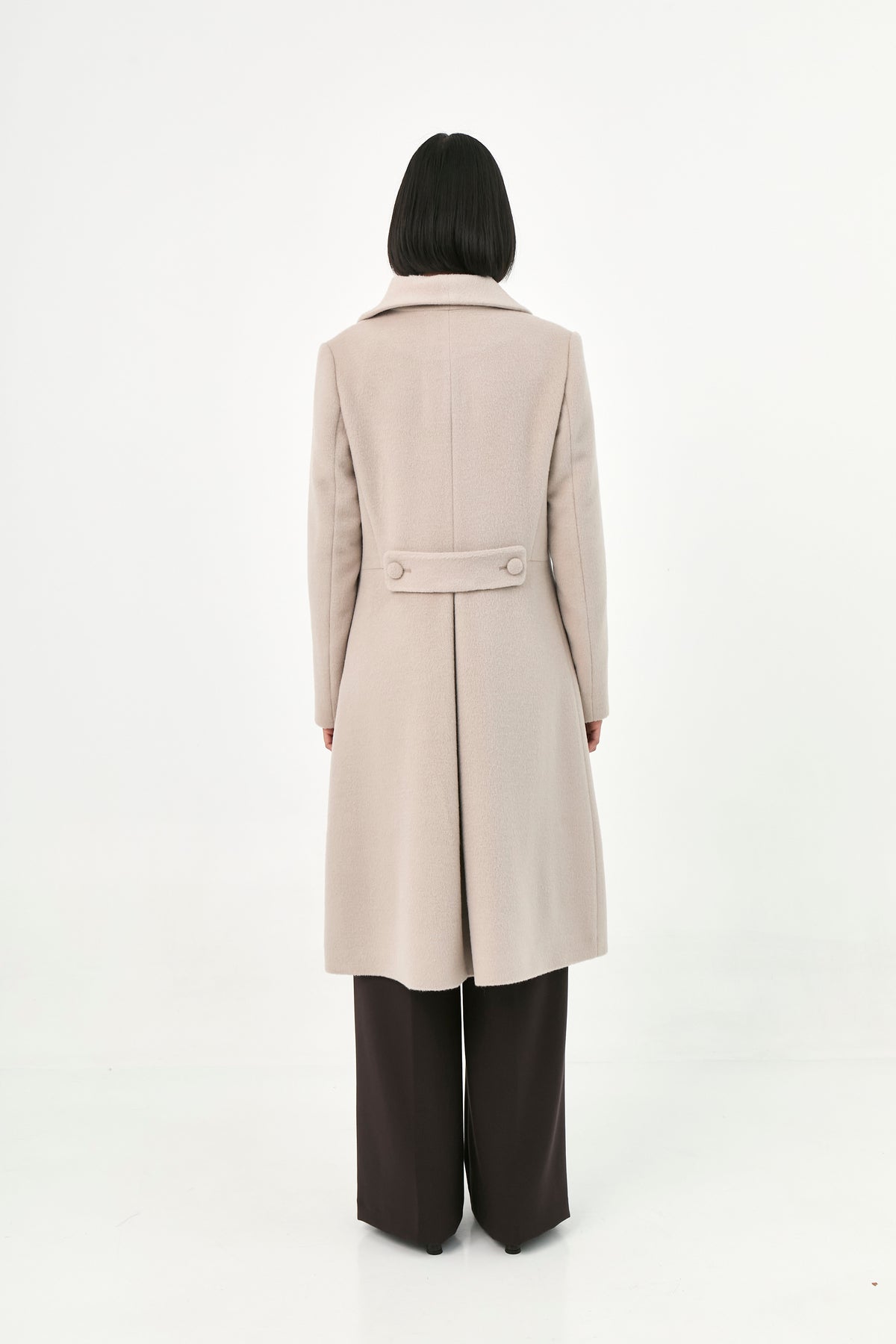 A model viewed from the back wearing a long, light beige baby alpaca coat with a martingale.