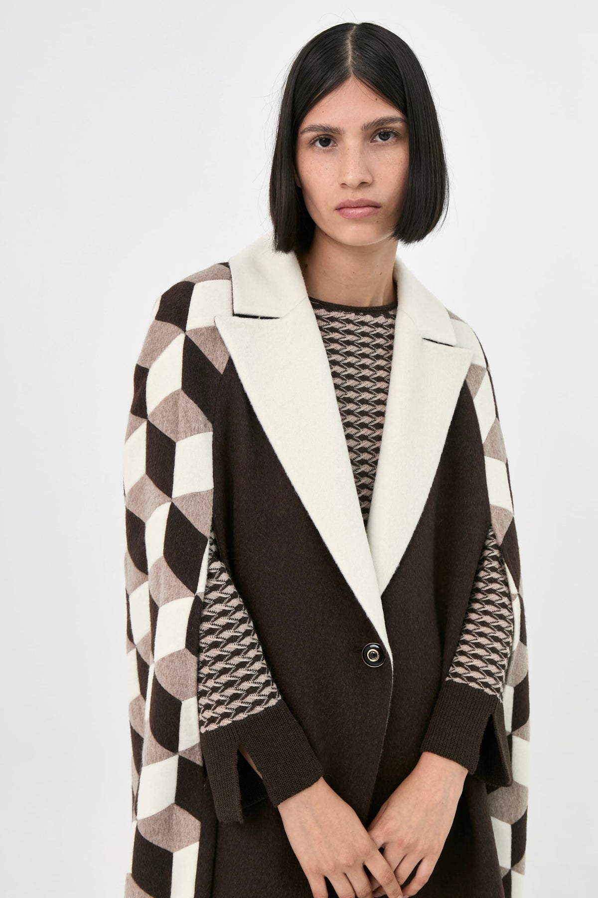 A model wearing a dark brown open-front cape coat with a thick cream collar and geometric patterned sleeves.