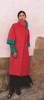 Person wearing a red coat with green accents standing against a stone wall.