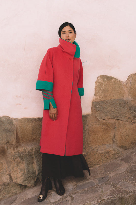 Person wearing a red coat with green accents standing against a stone wall.